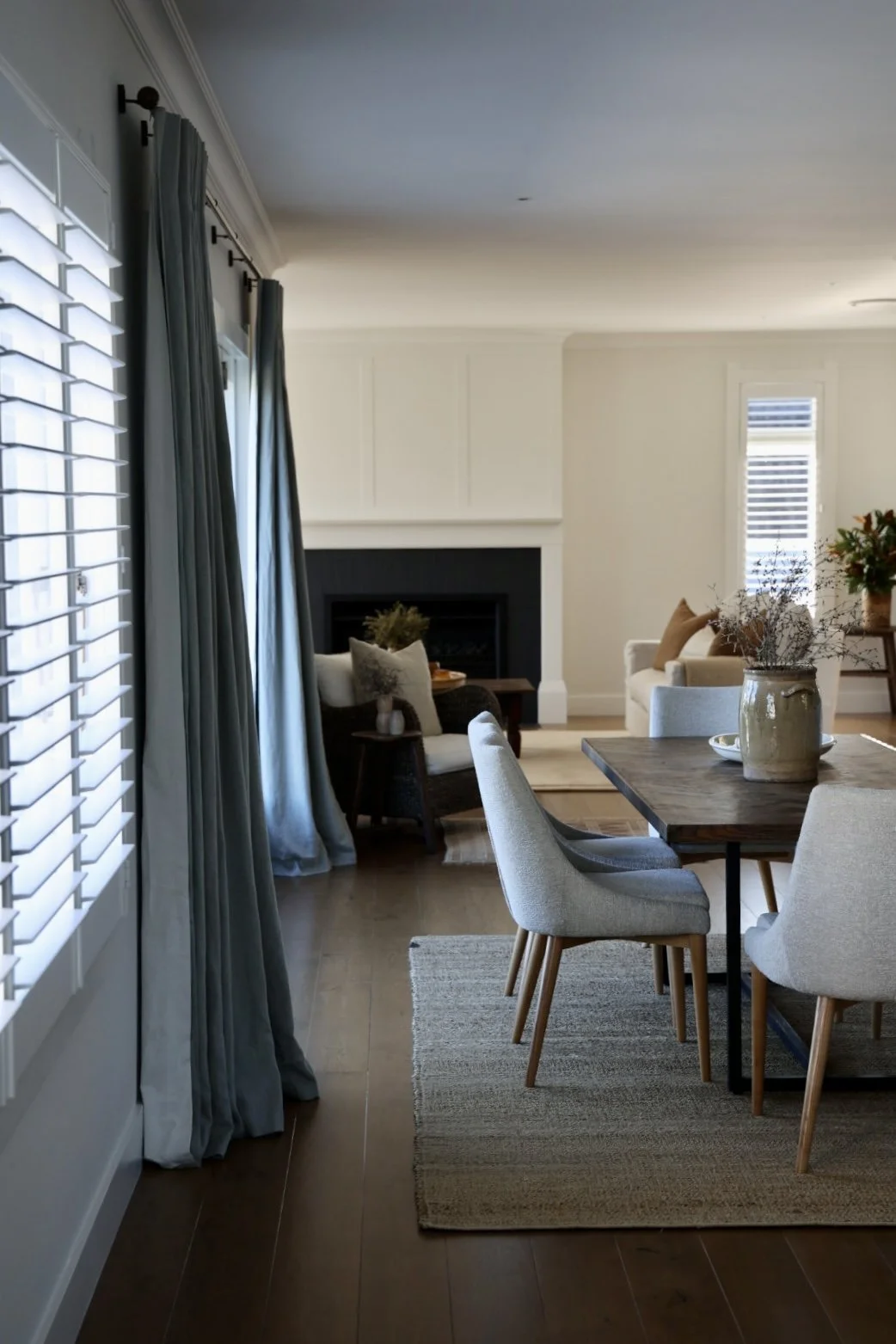 A cozy living room and dining area with light-colored walls, hardwood floors, and large windows with white plantation shutters and gray curtains. The space features a fireplace, a wooden dining table with cozy chairs, and decorative plants in vases.