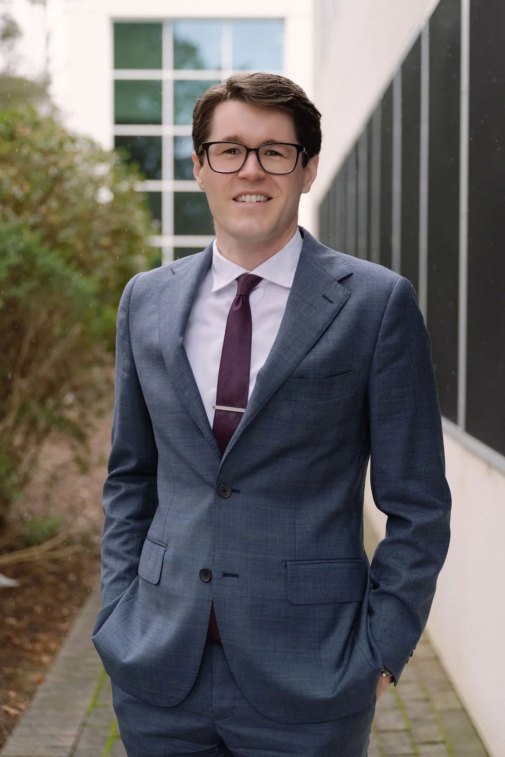 A young man wearing glasses and a gray business suit, standing outdoors in front of a modern building with large windows and greenery.