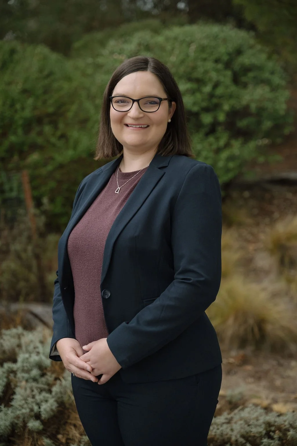 A woman in professional attire standing outdoors, smiling, with greenery and trees in the background.