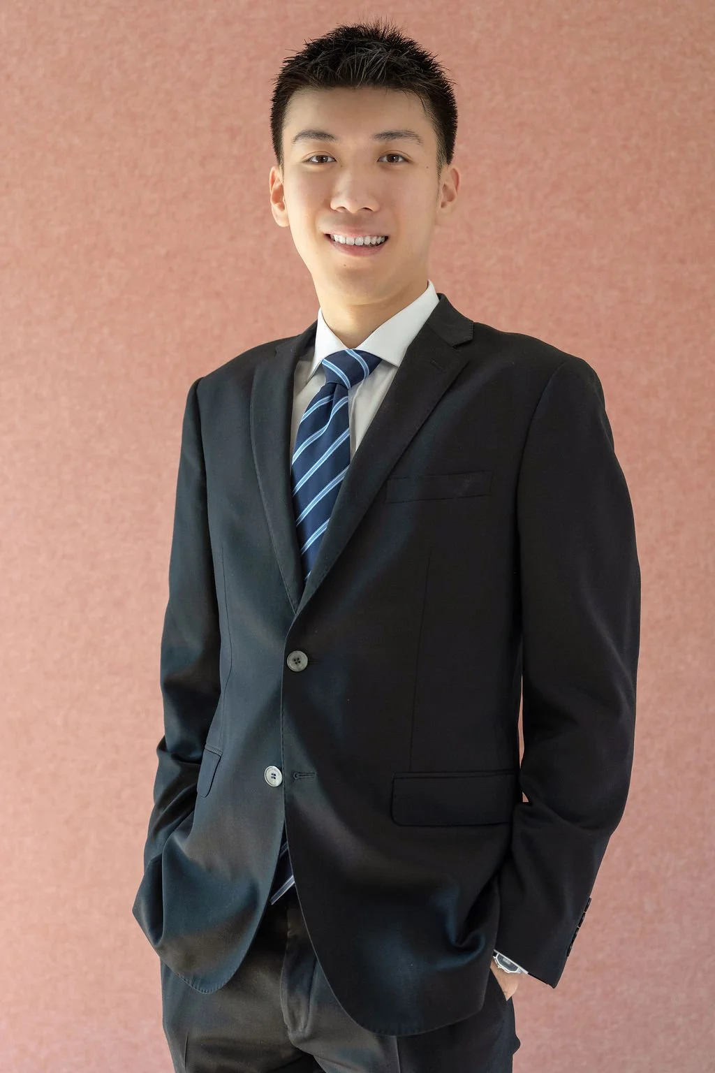 Young man in a black suit and blue striped tie smiling against a pinkish background.