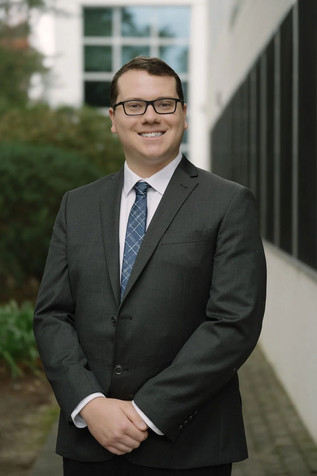 A smiling man in a dark suit, white shirt, and blue patterned tie standing outdoors with a building and greenery in the background.