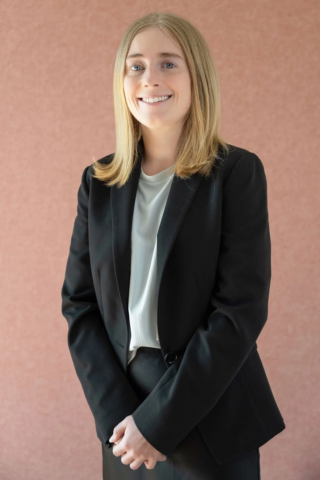 A young woman with shoulder-length blonde hair, wearing a black blazer and a white top, standing against a plain pinkish background, smiling at the camera.