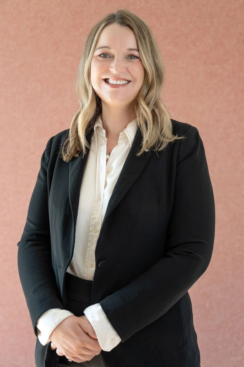 A smiling woman with shoulder-length light brown hair, wearing a black blazer over a white blouse, standing in front of a pink textured background.
