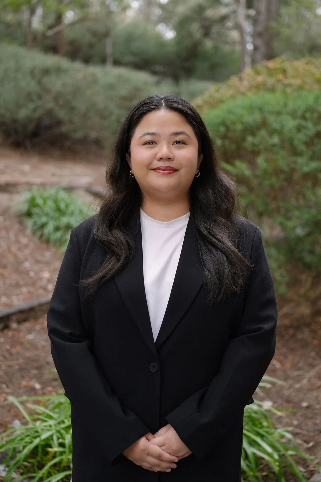 A woman with long dark hair wears a black blazer and white shirt, standing outdoors in a natural setting with trees and green bushes in the background.