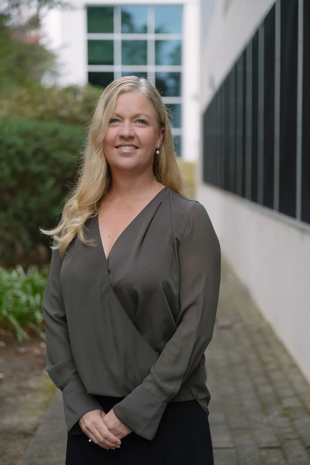 A woman with long blonde hair and pearl earrings standing outdoors on a paved walkway, wearing a dark gray blouse, with modern office buildings and greenery in the background.
