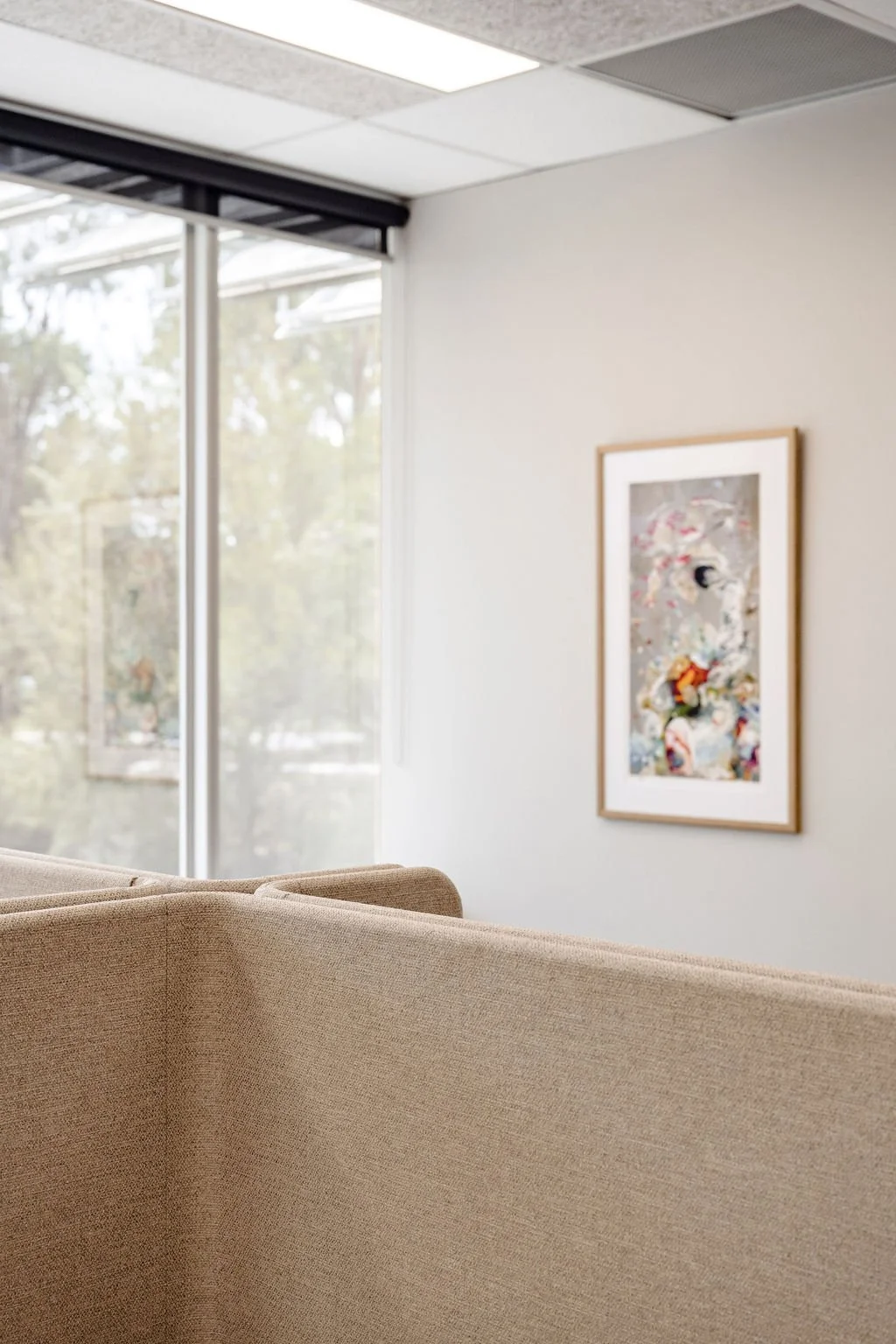 Interior of an office or waiting room with beige fabric cubicle walls, a large window with blinds, and framed artwork on a white wall.