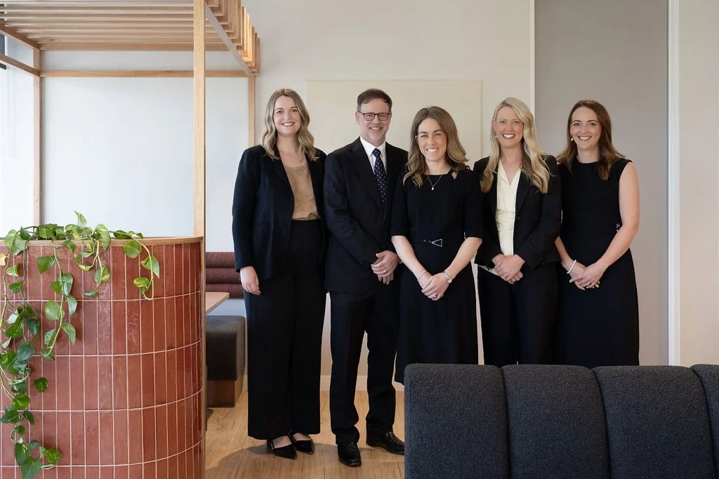 Group of five professionally dressed people standing indoors, smiling at the camera.
