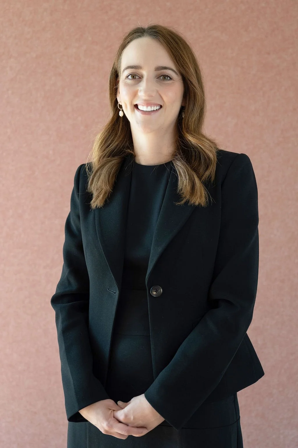 A woman with brown hair smiling, wearing a black blazer and earrings, standing in front of a pink textured wall.