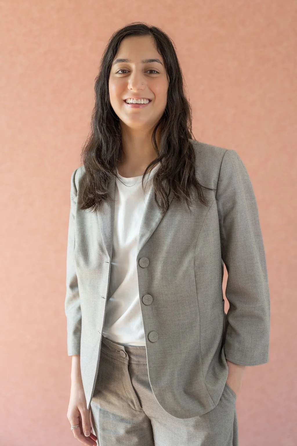 Young woman with dark, wavy hair wearing a light grey blazer and matching pants, smiling, standing against a soft pink background.