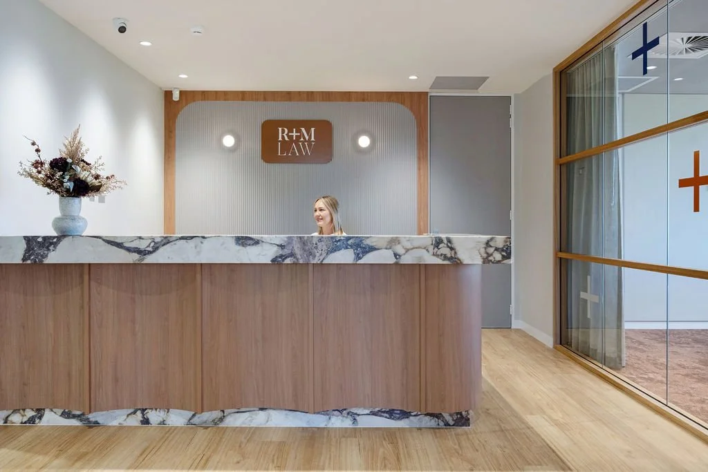 Reception desk at R+M Law with a marble top and wooden base, a woman standing behind the desk, a flower vase to the left, and glass walls on the right with colored crosses