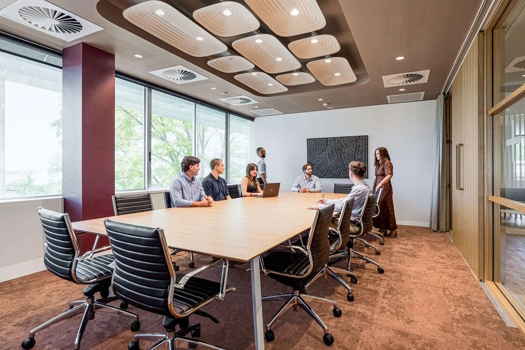 Business meeting in a modern conference room with ten people, some seated and some standing, with large window view and contemporary ceiling design.