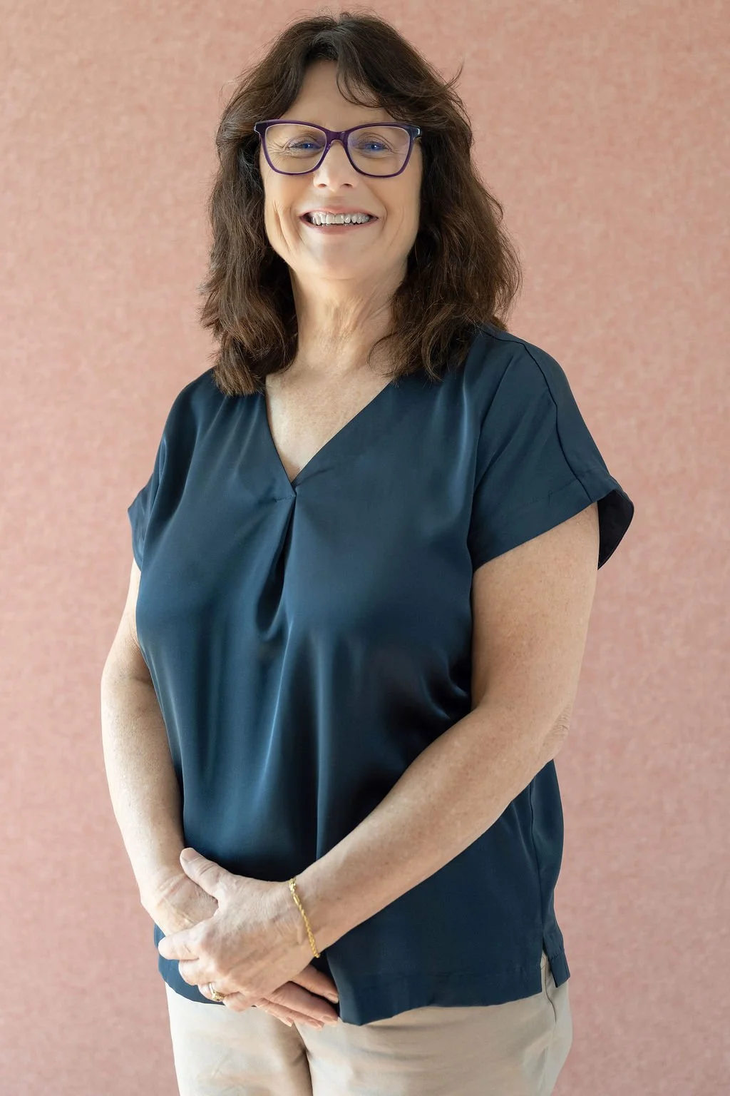 A middle-aged woman with shoulder-length brown hair, wearing glasses, a dark blue blouse, and khaki pants, standing against a textured pink wall and smiling at the camera.