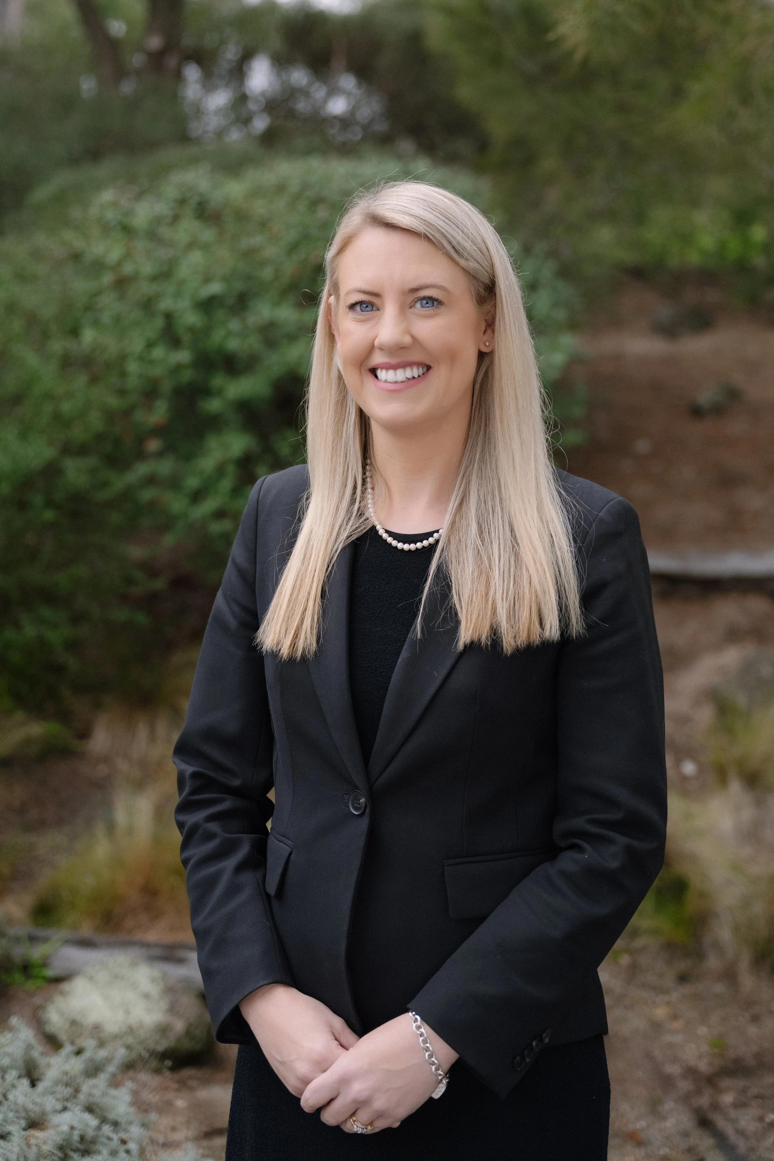 A woman with long blonde hair, dressed in a black blazer and black top, smiling outdoors with greenery in the background.
