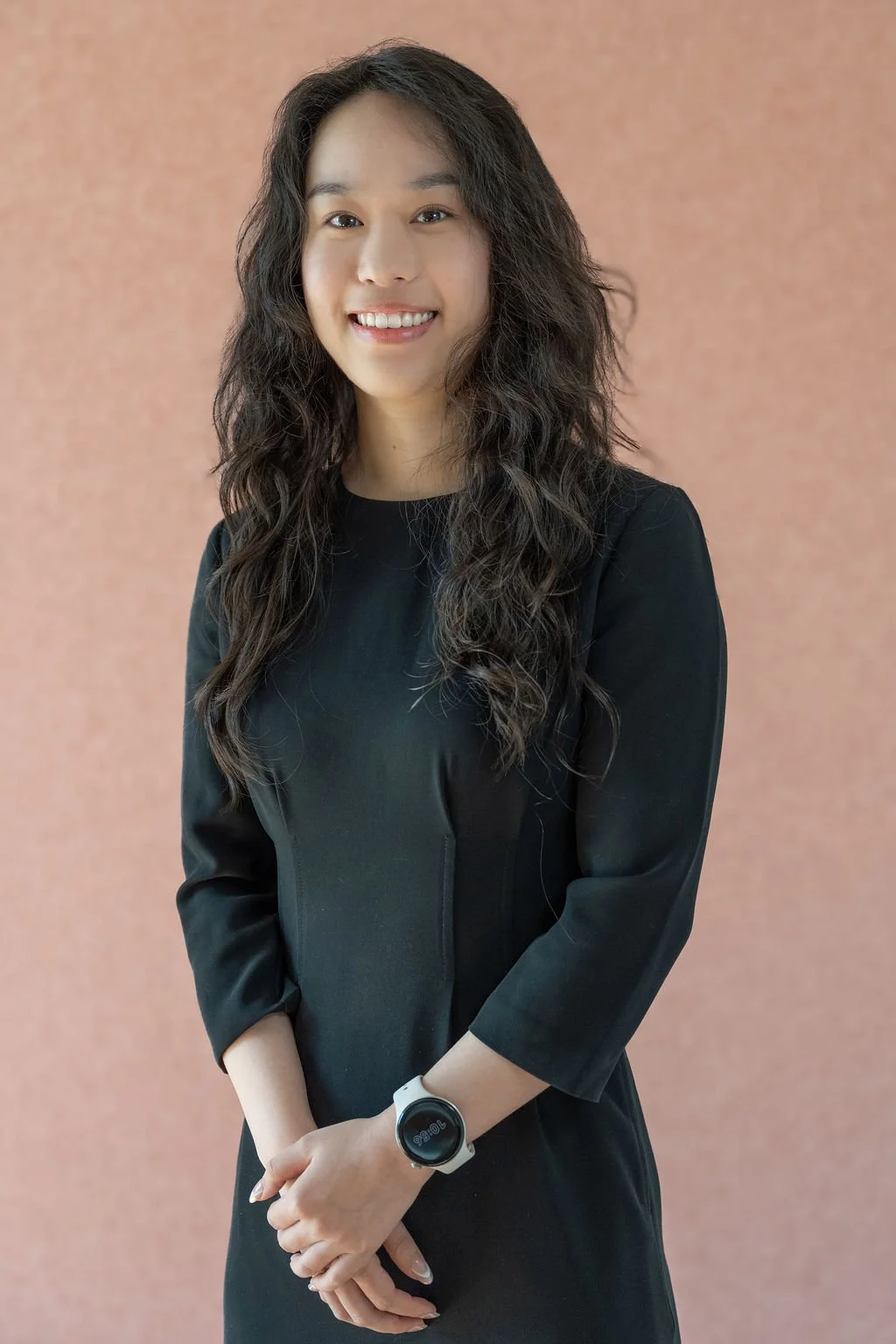 A smiling woman with long, wavy dark hair wearing a black dress and smartwatch, standing against a pinkish-beige background.