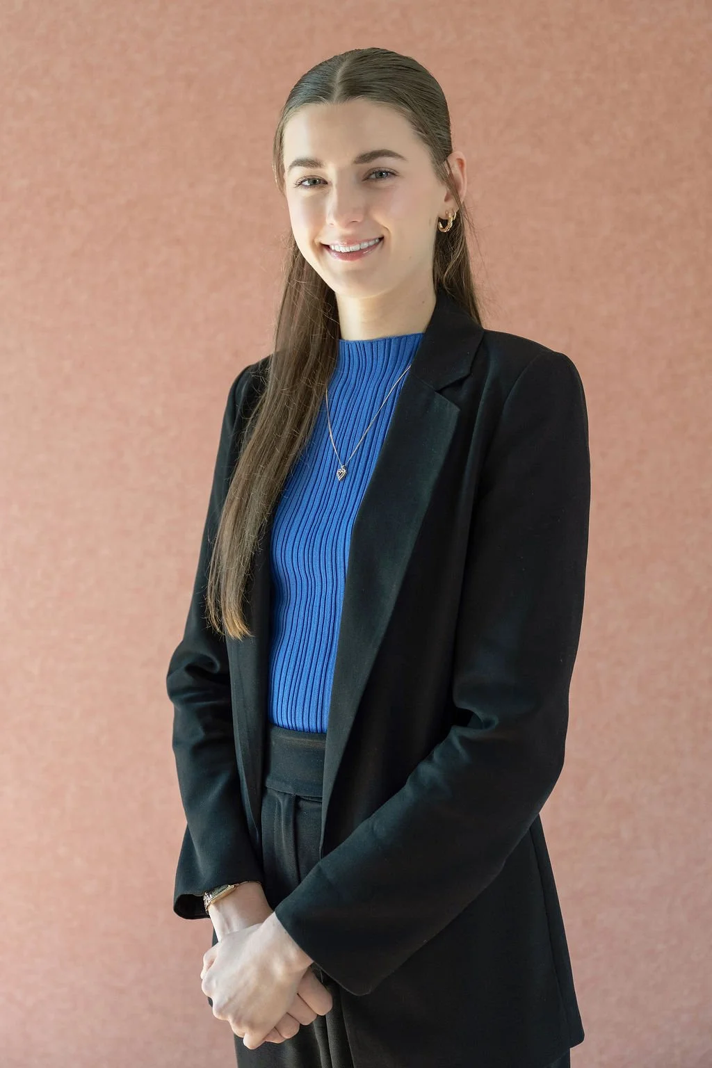 A young woman with long brown hair, smiling, wearing a black blazer and matching pants, with a blue ribbed top, necklaces, and earrings, standing in front of a mauve-colored background.