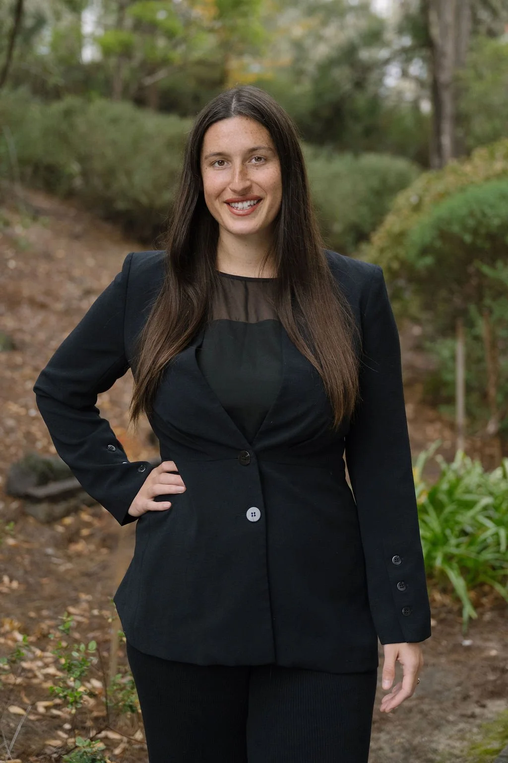 A woman with long dark hair, wearing a black blazer and top, stands outdoors with trees and bushes in the background, smiling at the camera.
