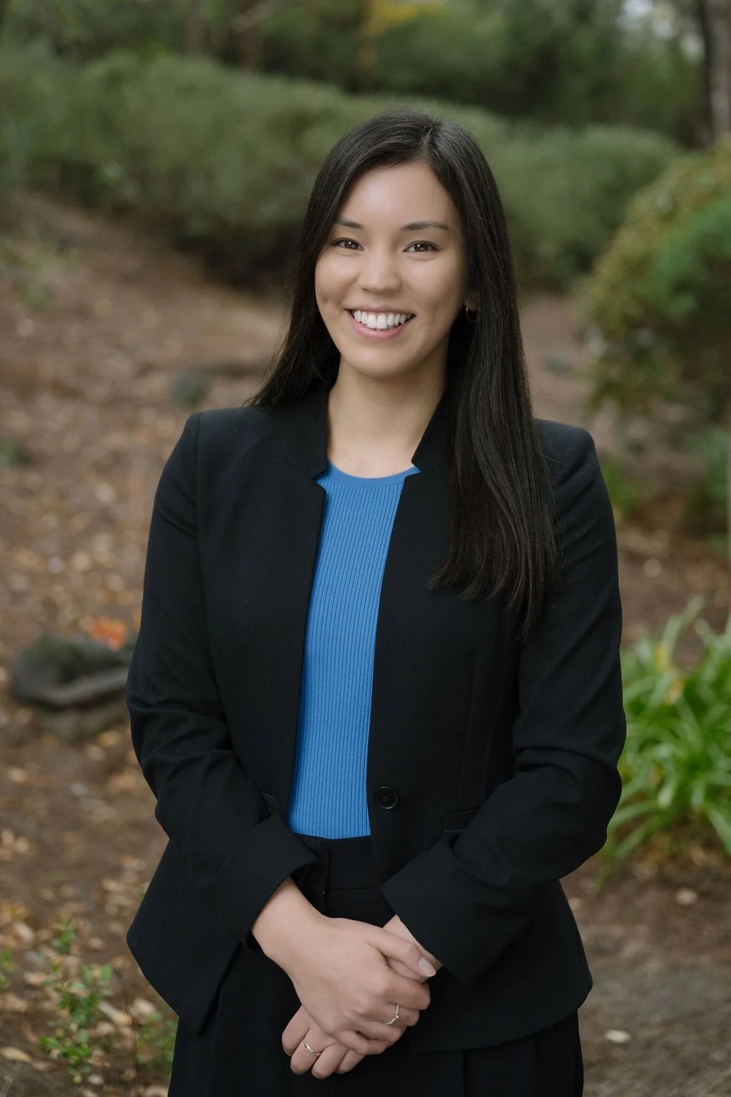 A woman with long black hair smiling outdoors, wearing a black blazer over a blue top, standing on a dirt path with green foliage in the background.