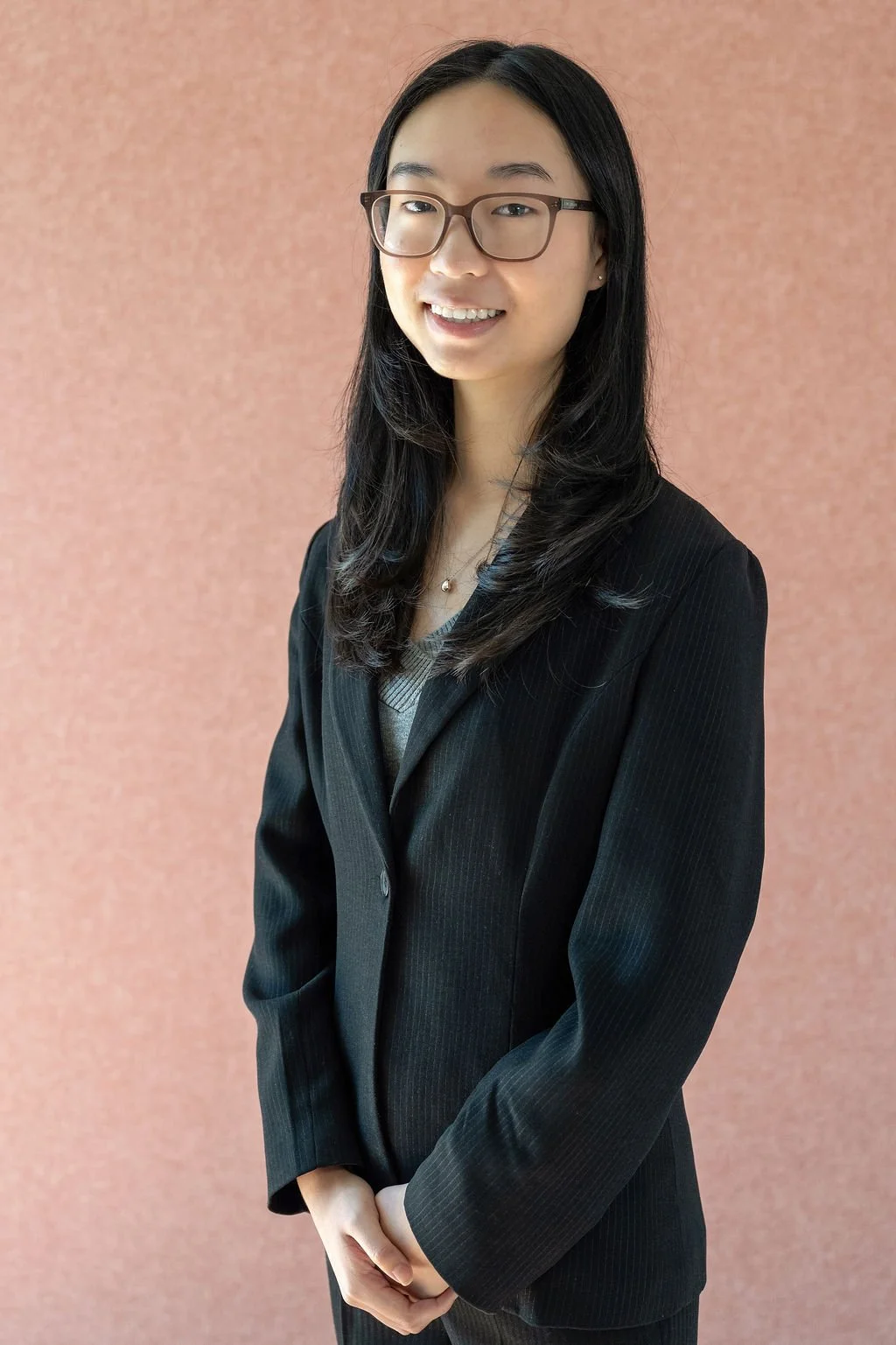 A young woman with long black hair and glasses smiling, dressed in a black blazer and gray shirt, standing against a pink indoor wall.
