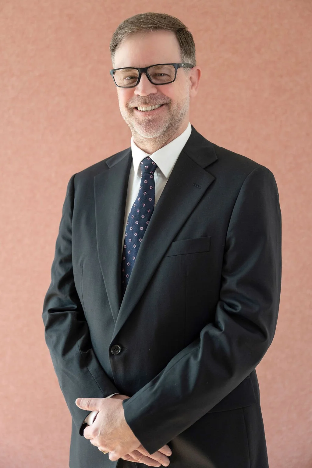 A middle-aged man with glasses, wearing a black suit, white shirt, and patterned tie, smiling and standing against a plain, textured peach-colored background.