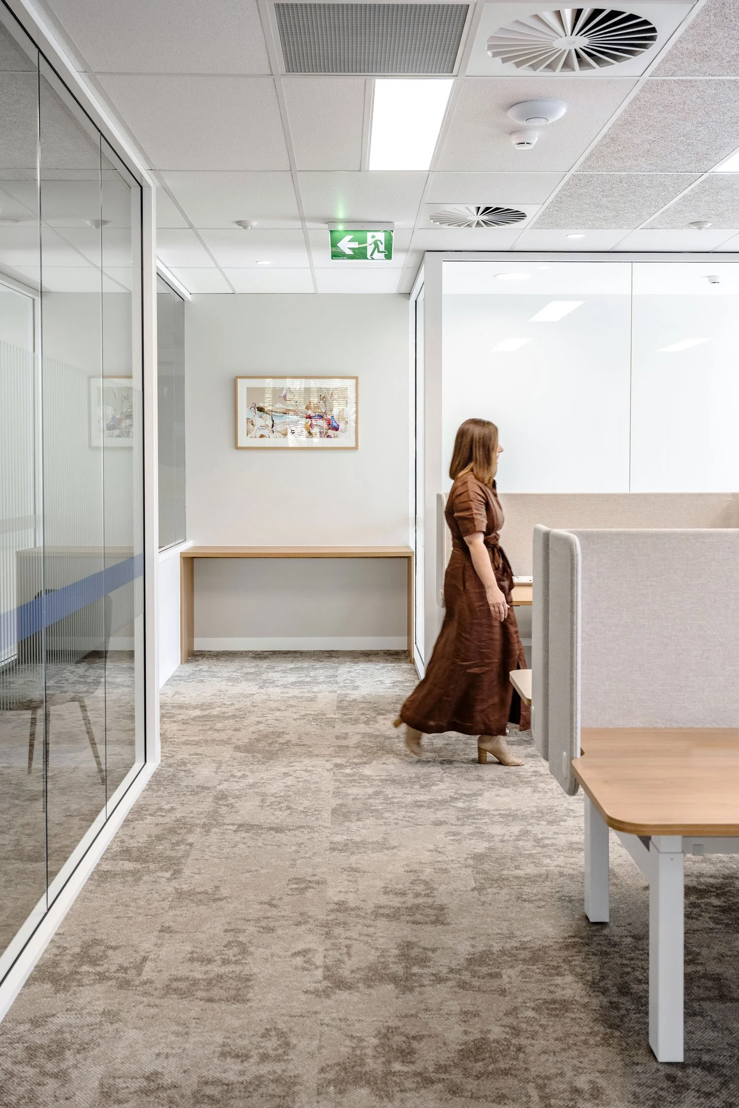 An office hallway with beige carpet, white walls, and ceiling tiles. A woman in a brown dress and beige heeled boots is walking past a cubicle wall. There are framed pictures on the wall, a wooden table with a beige top, and an emergency exit sign on the ceiling.