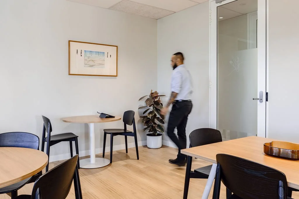 An office or meeting room with a man walking near a large potted plant. The room has white walls, wooden flooring, and a glass door. There are round tables with black and wood chairs, some with a small tray or phone on the table.