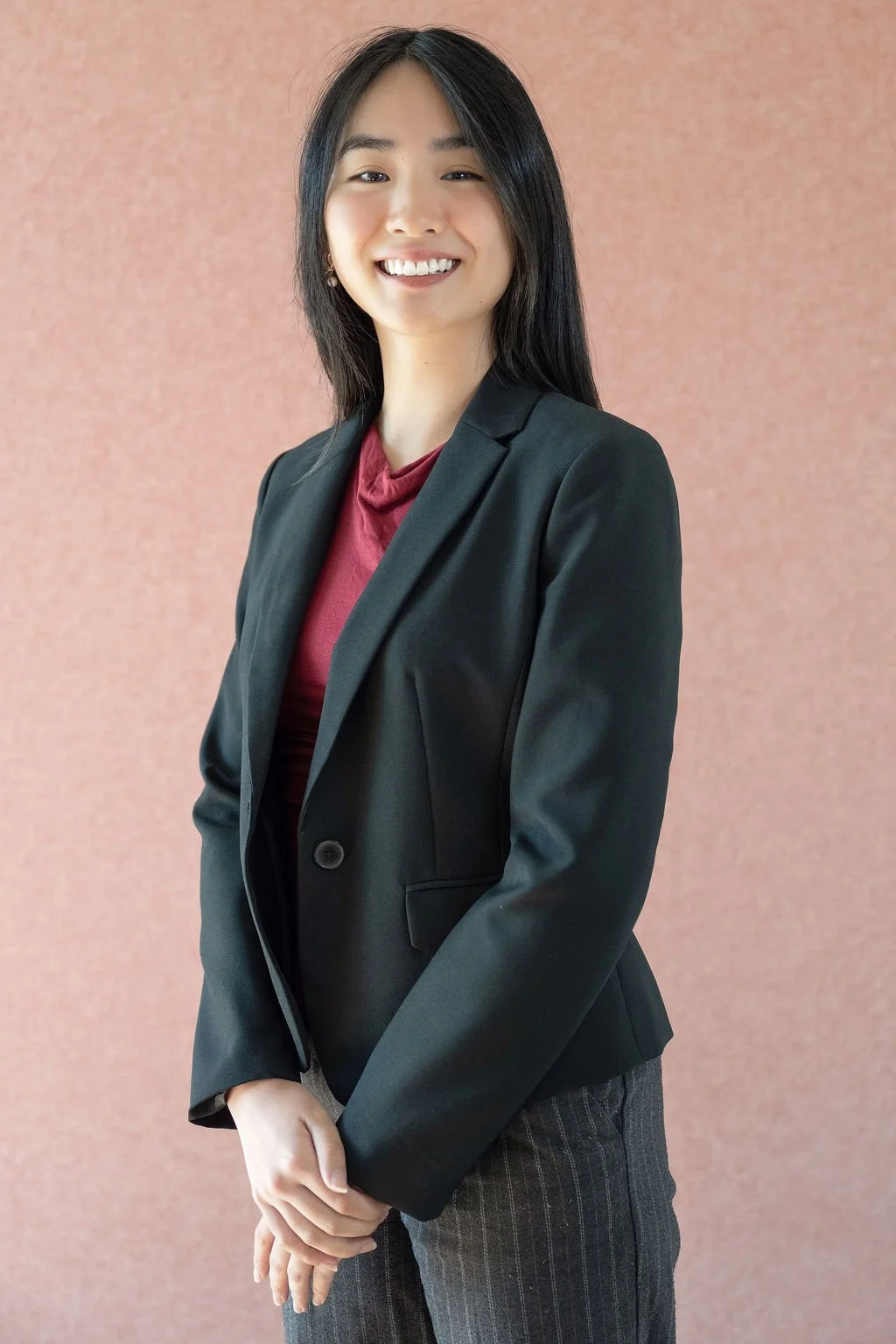 A young woman with straight black hair wearing a black blazer over a dark red top, posing against a pinkish textured background, smiling with her hands clasped in front of her.