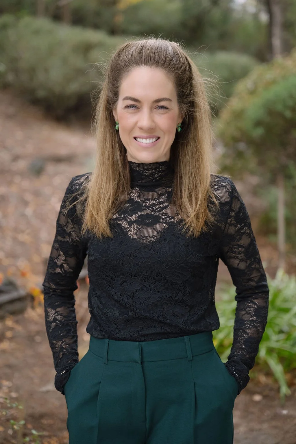 A woman standing outdoors on a trail, wearing a black lace top and green pants, smiling at the camera.