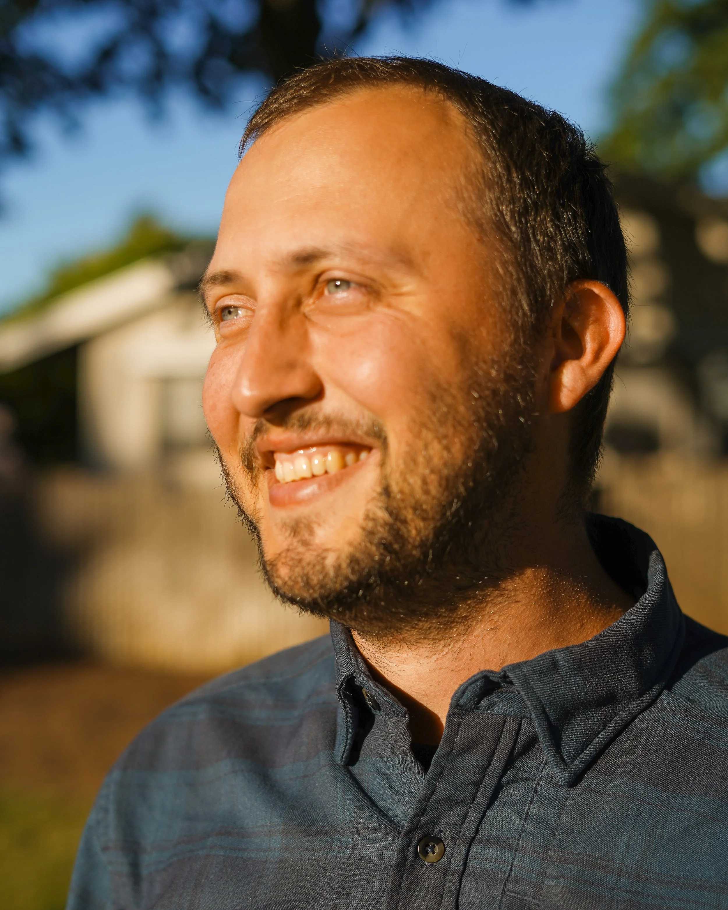 Close-up of smiling man with short dark hair, beard, and light-colored eyes, wearing a dark collared shirt, outdoors on sunny day.