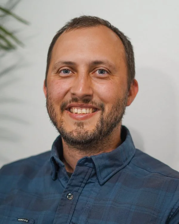 A smiling man with short brown hair, a beard, wearing a blue button-up shirt, against a plain light background.