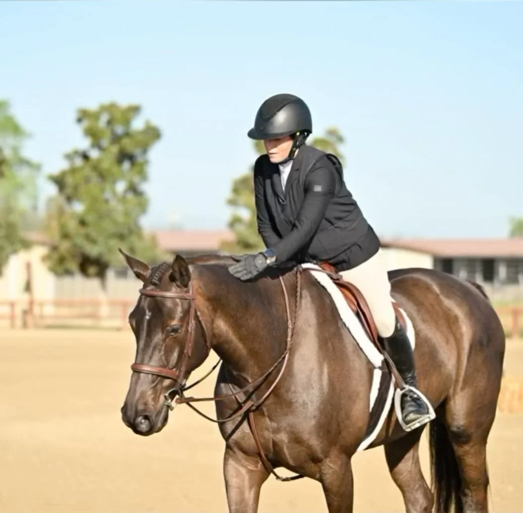 A woman wearing a black riding helmet, black jacket, and white pants riding a brown horse in an outdoor arena with trees and a building in the background.