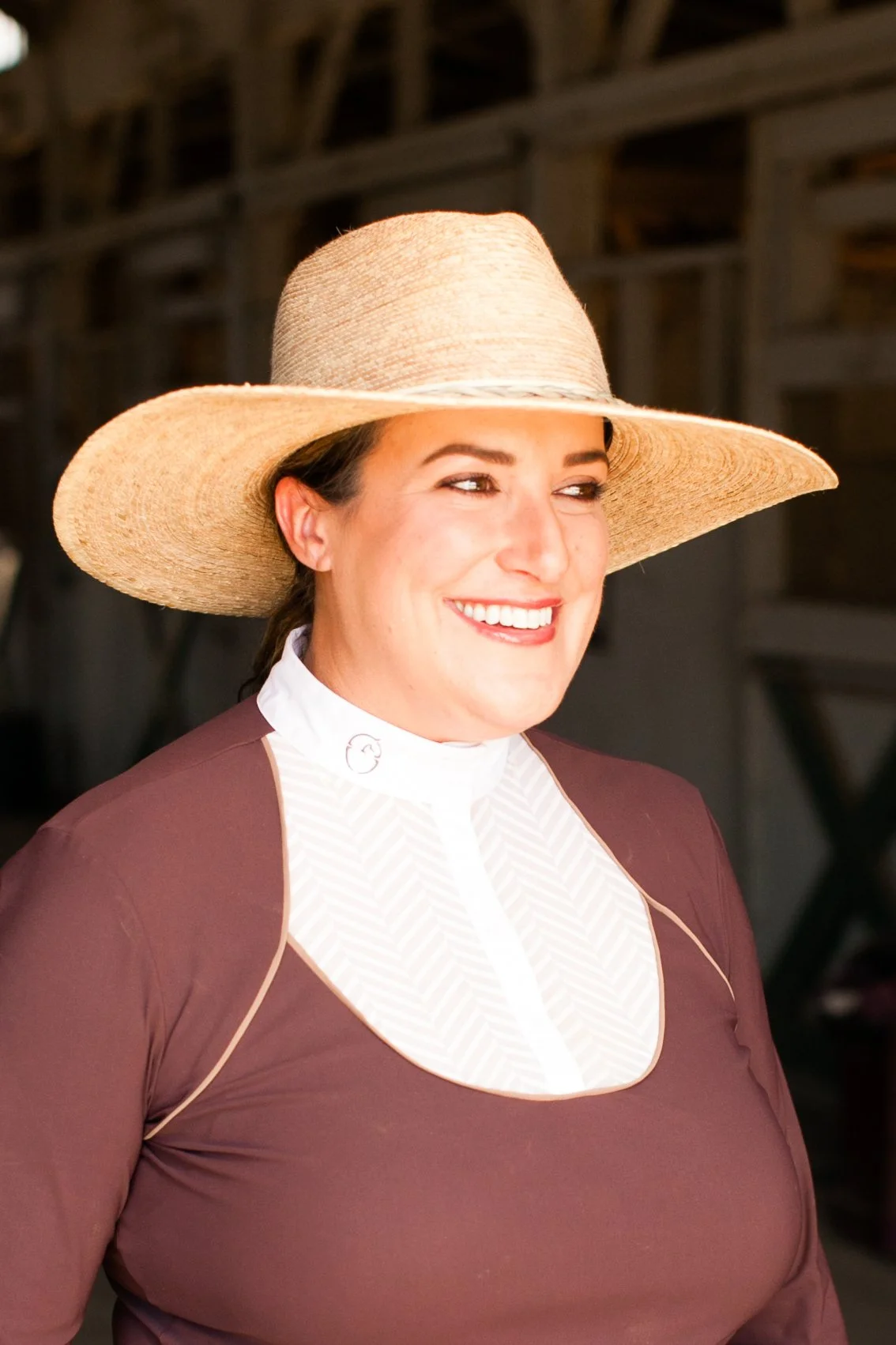 A smiling woman wearing a wide-brimmed straw hat and a brown top with a white patterned collar.