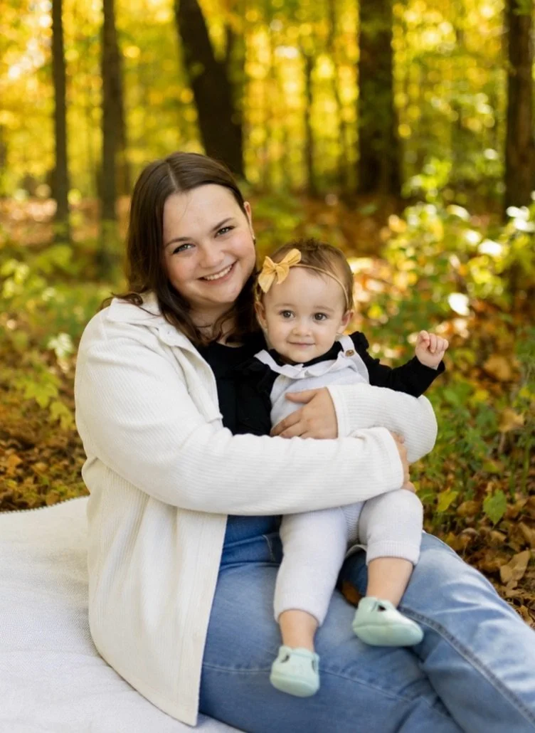 A woman and a young girl sitting in a forest with yellow and green foliage, smiling at the camera. The woman is holding the girl, who has a bow in her hair.
