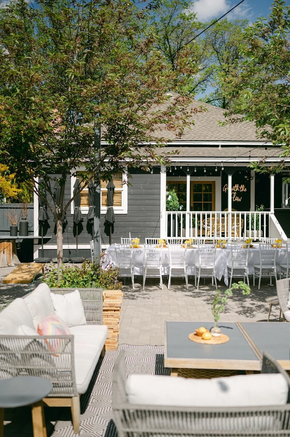 Outdoor patio setup with white chairs and tables for a gathering, surrounded by trees and greenery, with a house featuring a porch in the background.