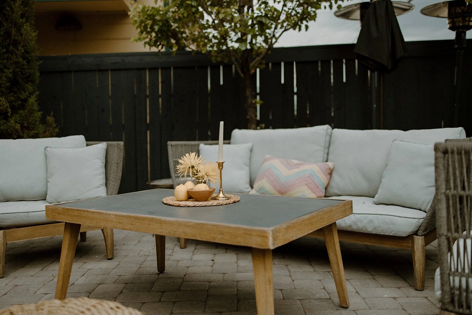 Outdoor patio with white cushioned sectional sofa, a wooden coffee table with a candle, floral decoration, and fruit bowls, surrounded by a black fence, a tree, and patio umbrellas in the background.