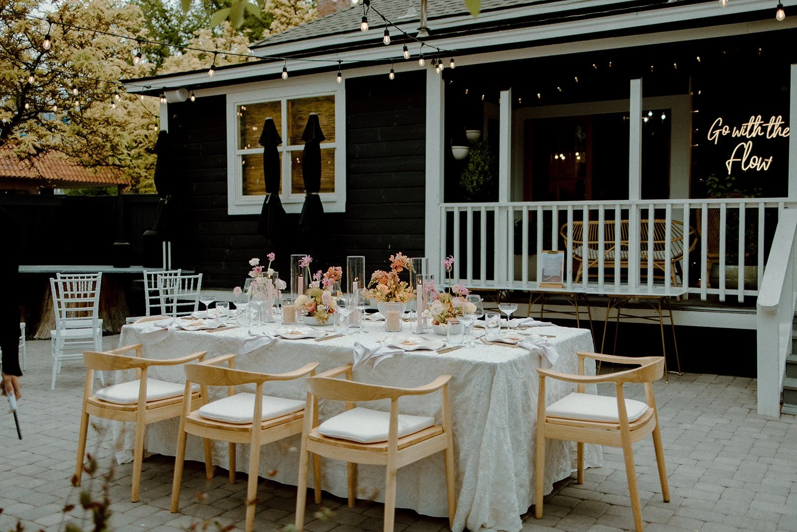 Outdoor dining area with a long table covered with a white tablecloth, decorated with pink and white flowers in vases, candles, and glassware. Wooden and white chairs surround the table. The setting appears ready for a celebration or event, with string lights overhead and a dark house or building in the background featuring a porch with a bench and a sign that reads 'Go with the flow'.