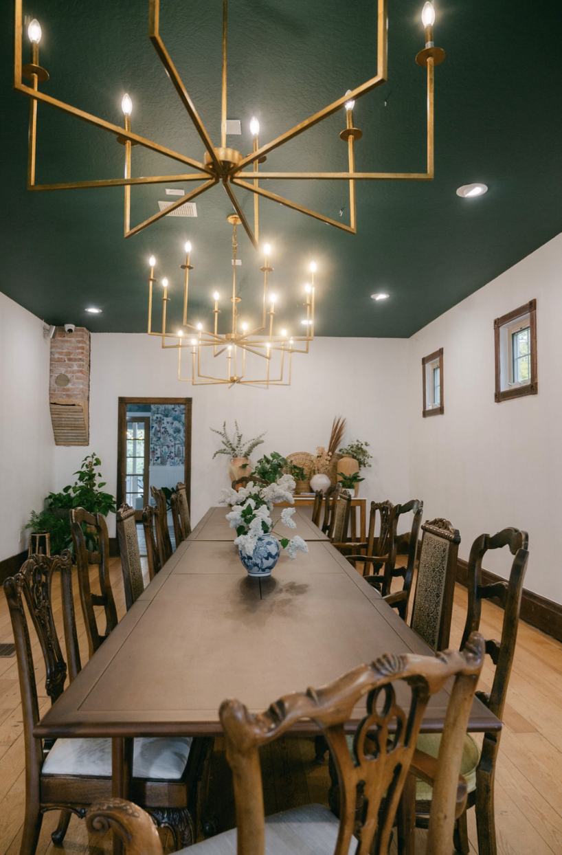 A long dining table with ornate wooden chairs, decorative vases with white flowers, and a green ceiling with two large modern chandeliers with candles. The room has white walls, small framed windows, and a door leading to another room with patterned 