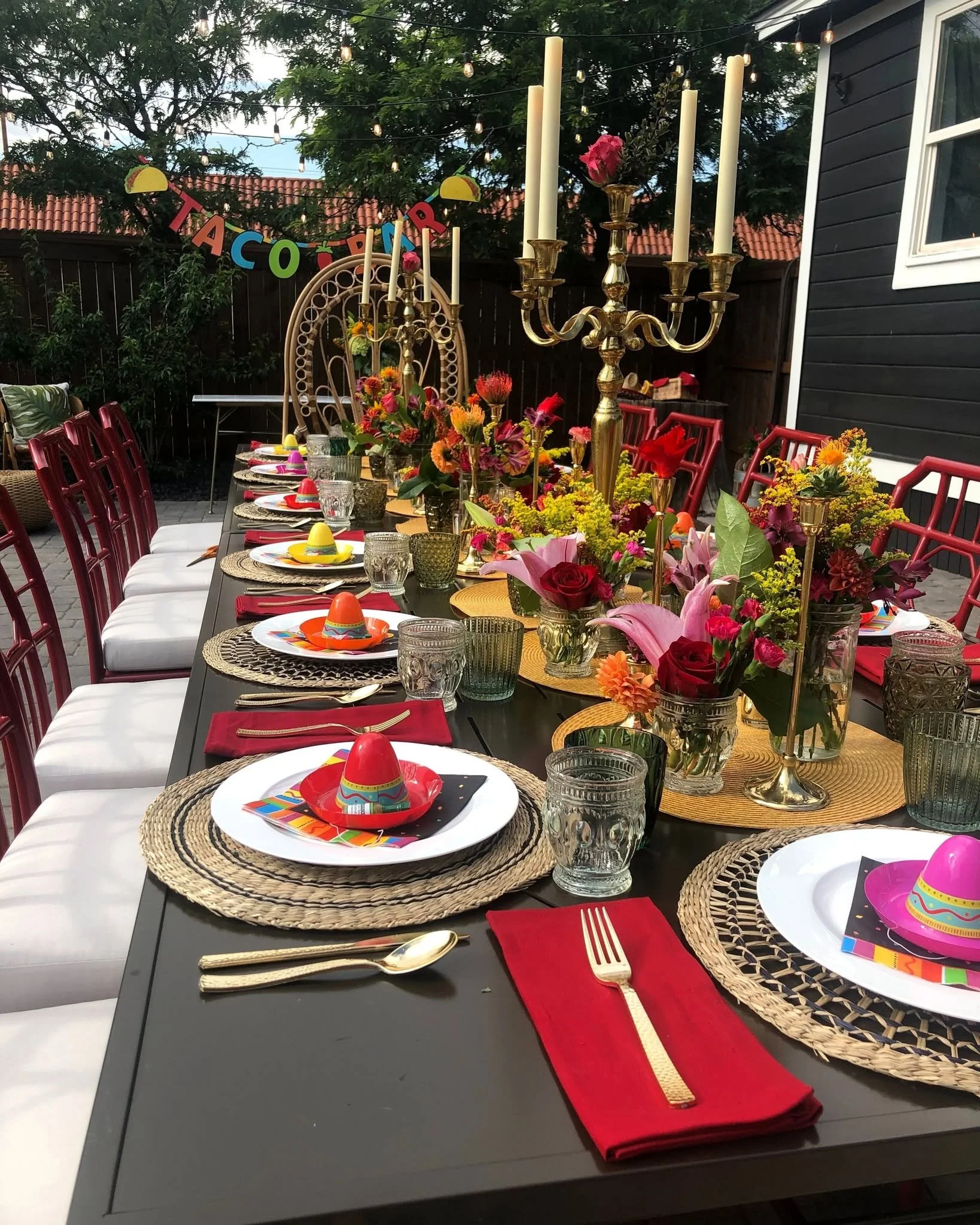 Outdoor dining table decorated for a celebration, with colorful floral arrangements, candles, party hats, and place settings including gold utensils, clear and colored glasses, and red napkins.