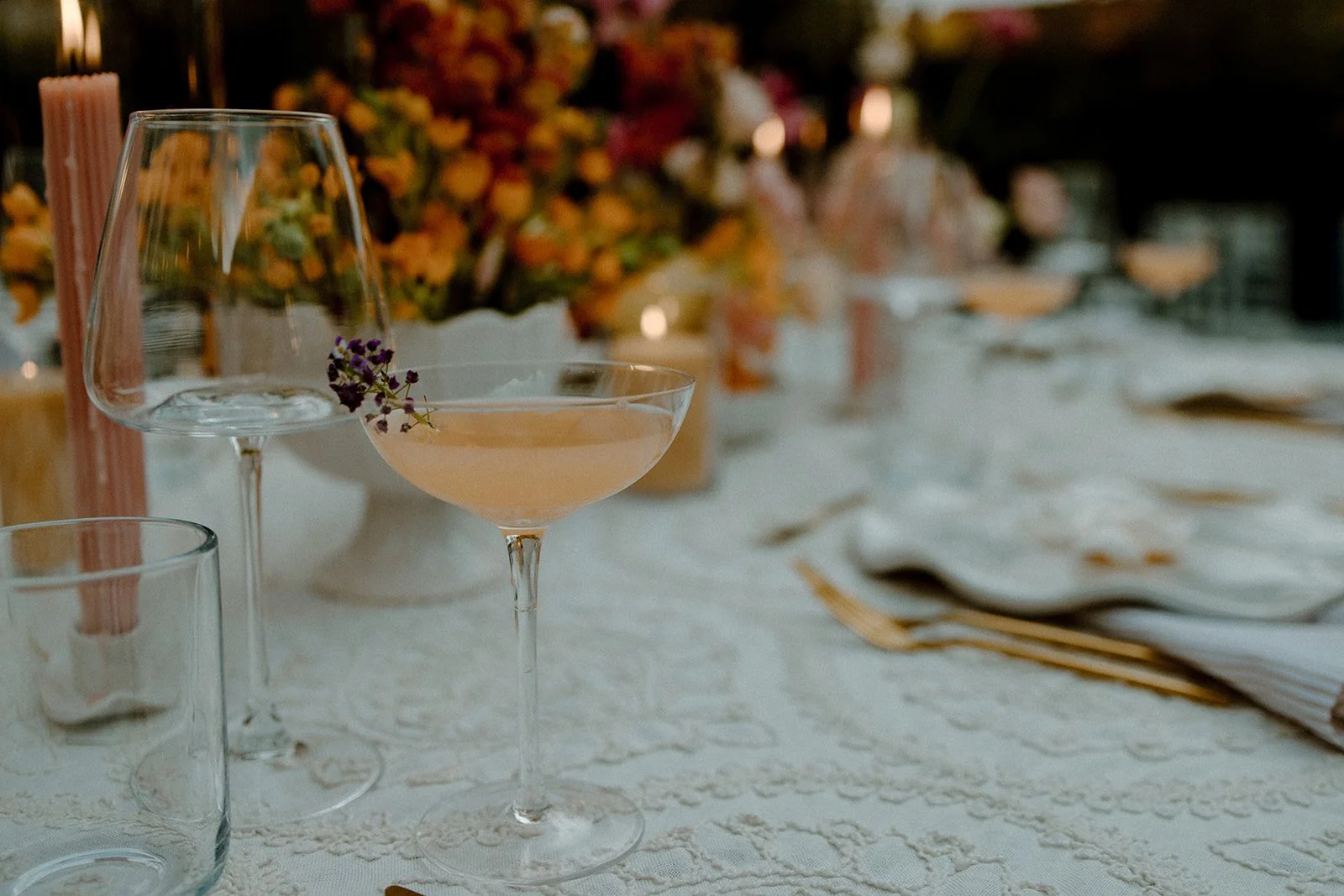 Close-up of stemware and cocktails on a set table with flowers and candles in the background.