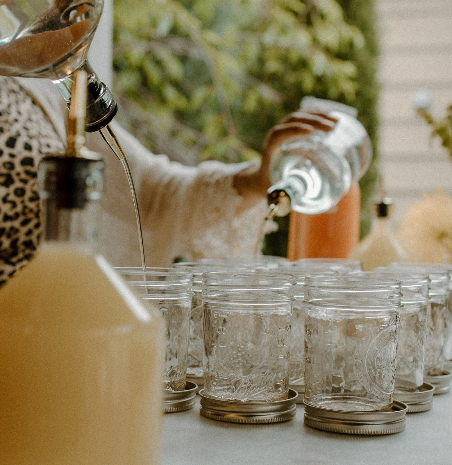 A person is pouring a beverage into clear glass jars arranged on a table, with several jars already filled or lined up. The person is holding a bottle with a napkin around the neck, using a dropper or spout to fill the jars. The background shows some