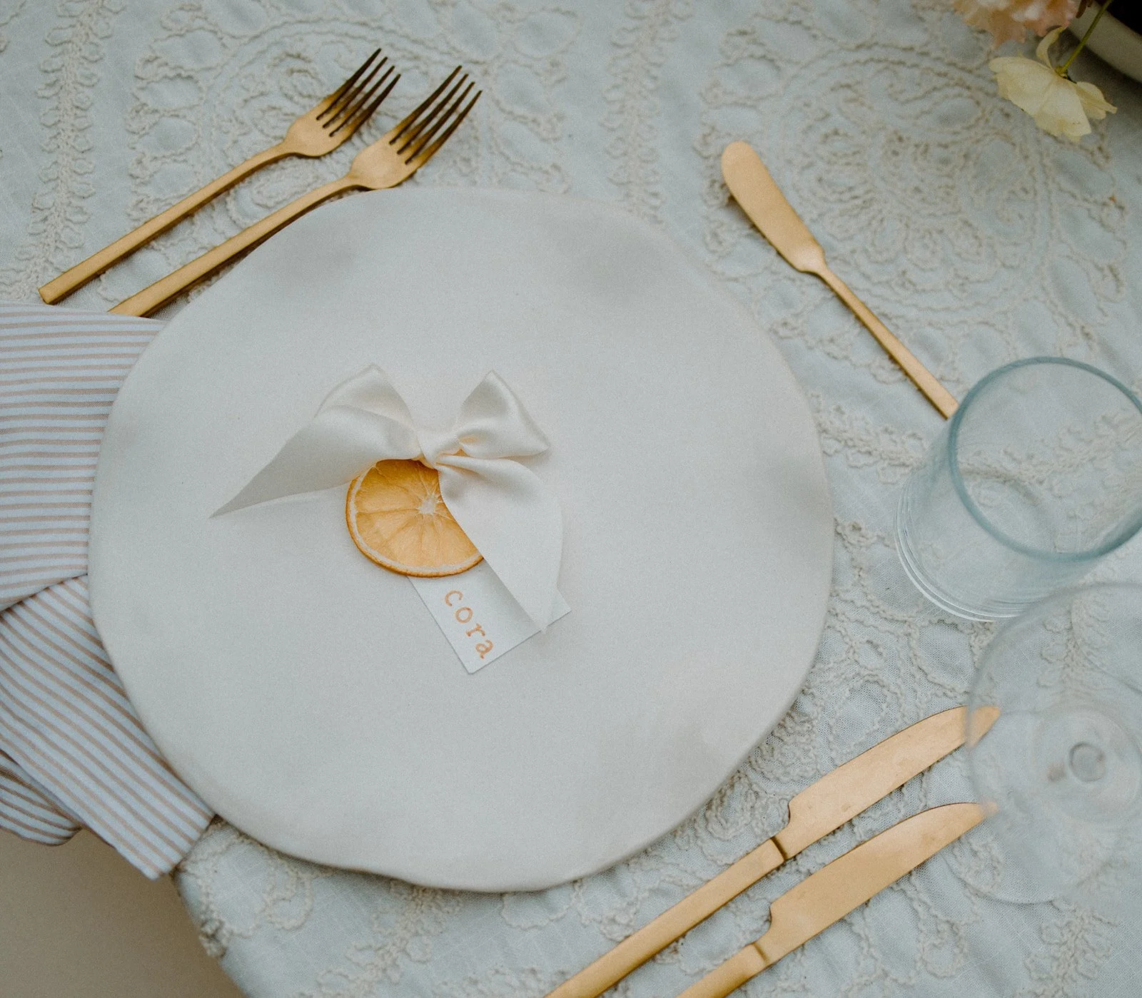 Elegant table setting with gold cutlery, a glass, flowers, and a white plate with a small orange slice and white ribbon decorated with a star and the word 'CORA' on a lace tablecloth.