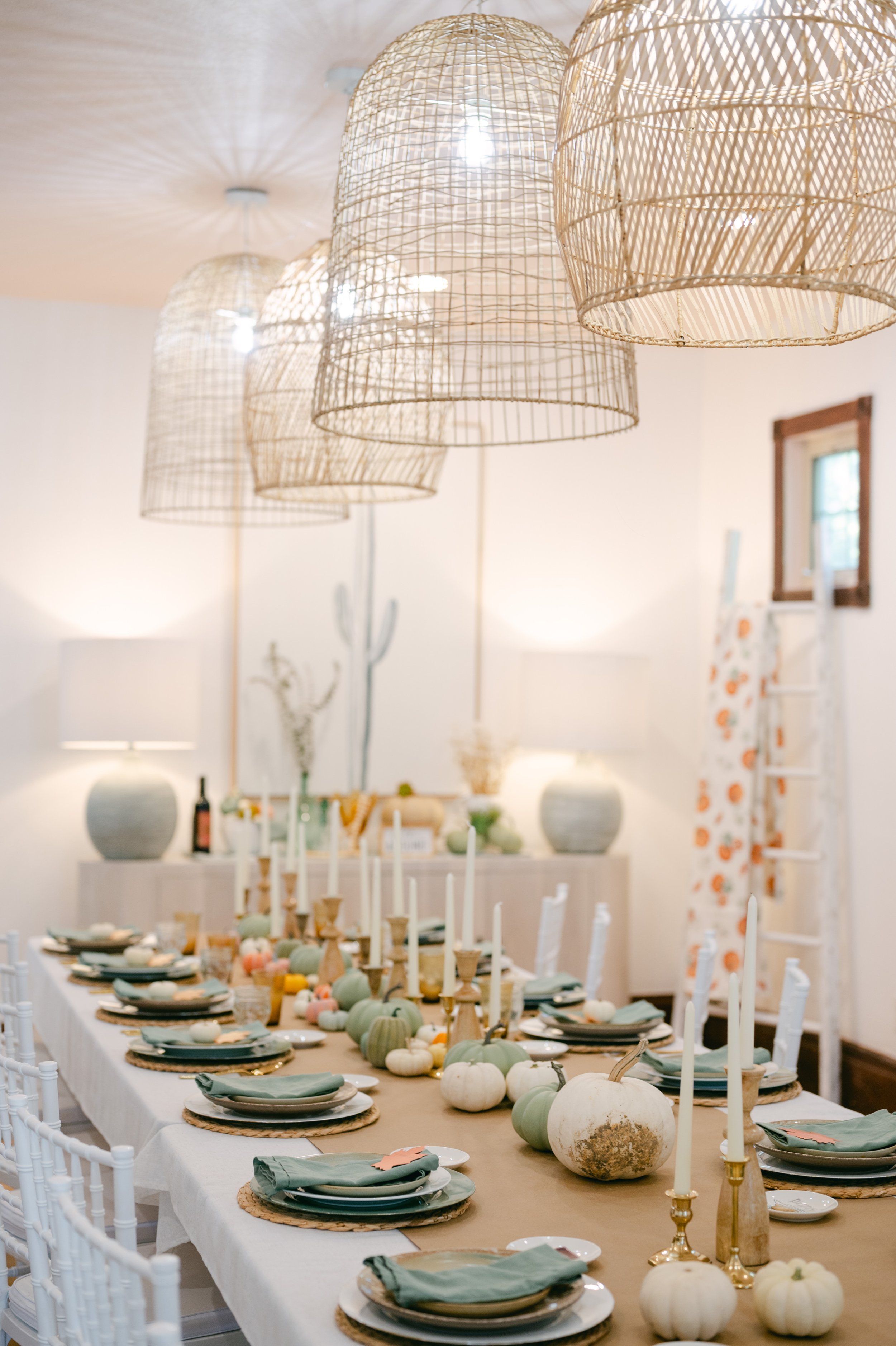 A dining table decorated with white and pastel pumpkins, candles, and place settings, with woven pendant lights hanging above in a bright room.