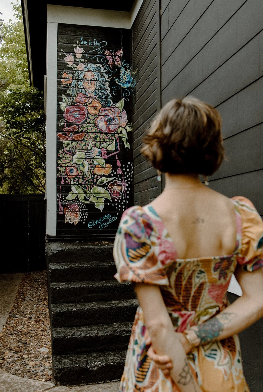 A woman with short curly hair wearing a colorful dress with floral and butterfly patterns is looking at a large colorful mural of flowers, a woman's face with flowing hair, and butterflies painted on a dark wall. The mural includes the text '@ivy.ase