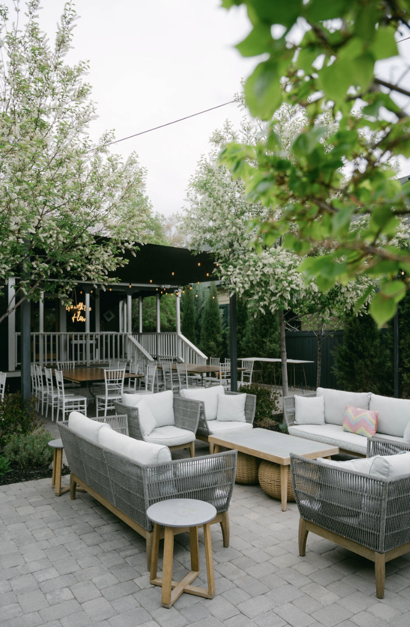 Outdoor patio with white cushioned wicker furniture, a wooden coffee table, and small round side tables. There are flowering trees and string lights overhead, creating a cozy, inviting atmosphere.