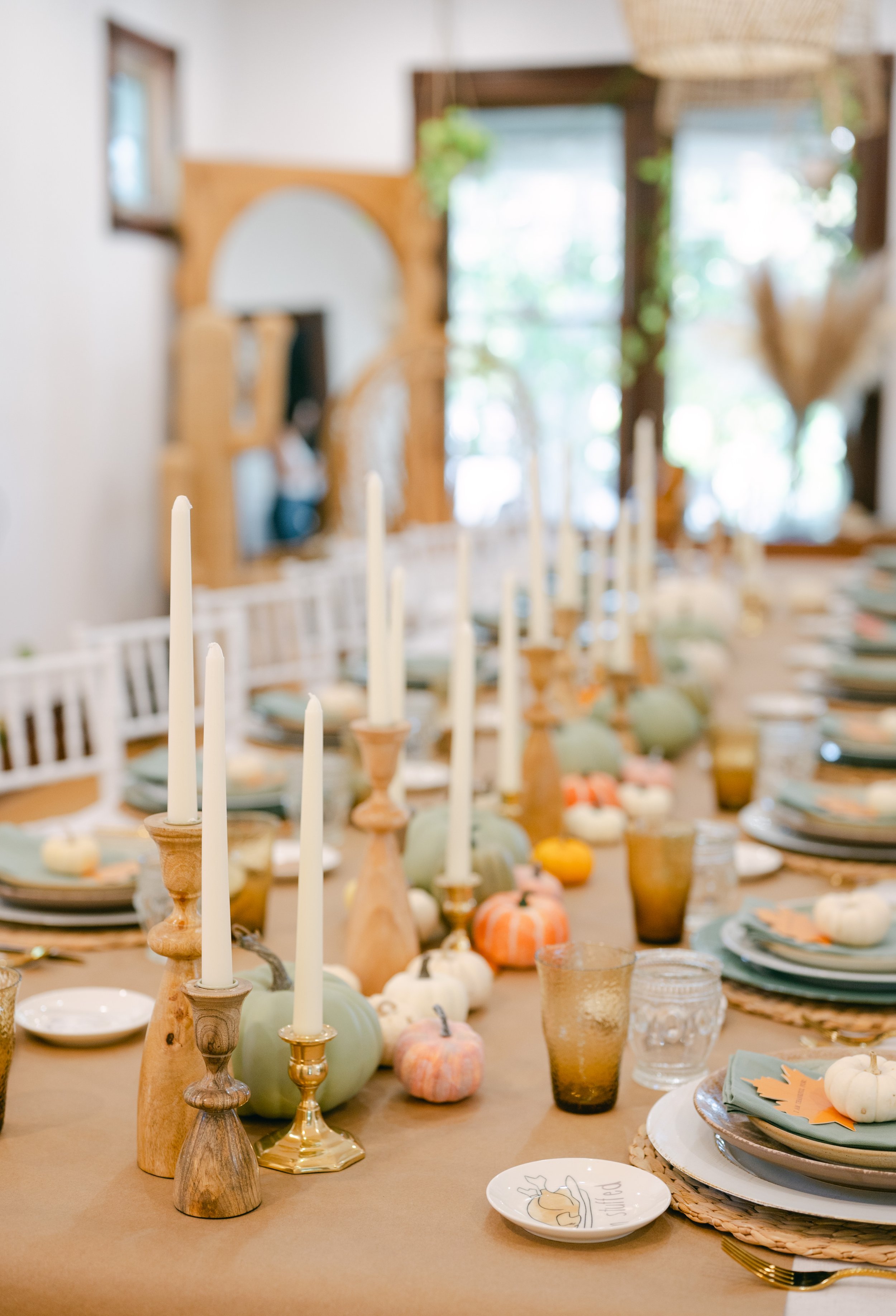 A rustic dining table set with fall-themed decorations including small pumpkins, candles, and place settings with napkins and plates in a bright room with large windows.