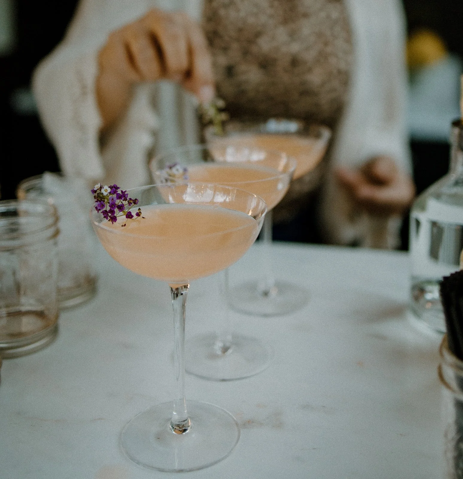 Three pink cocktails in coupe glasses garnished with purple and white flowers, set on a white table with jars and bottles in the background.