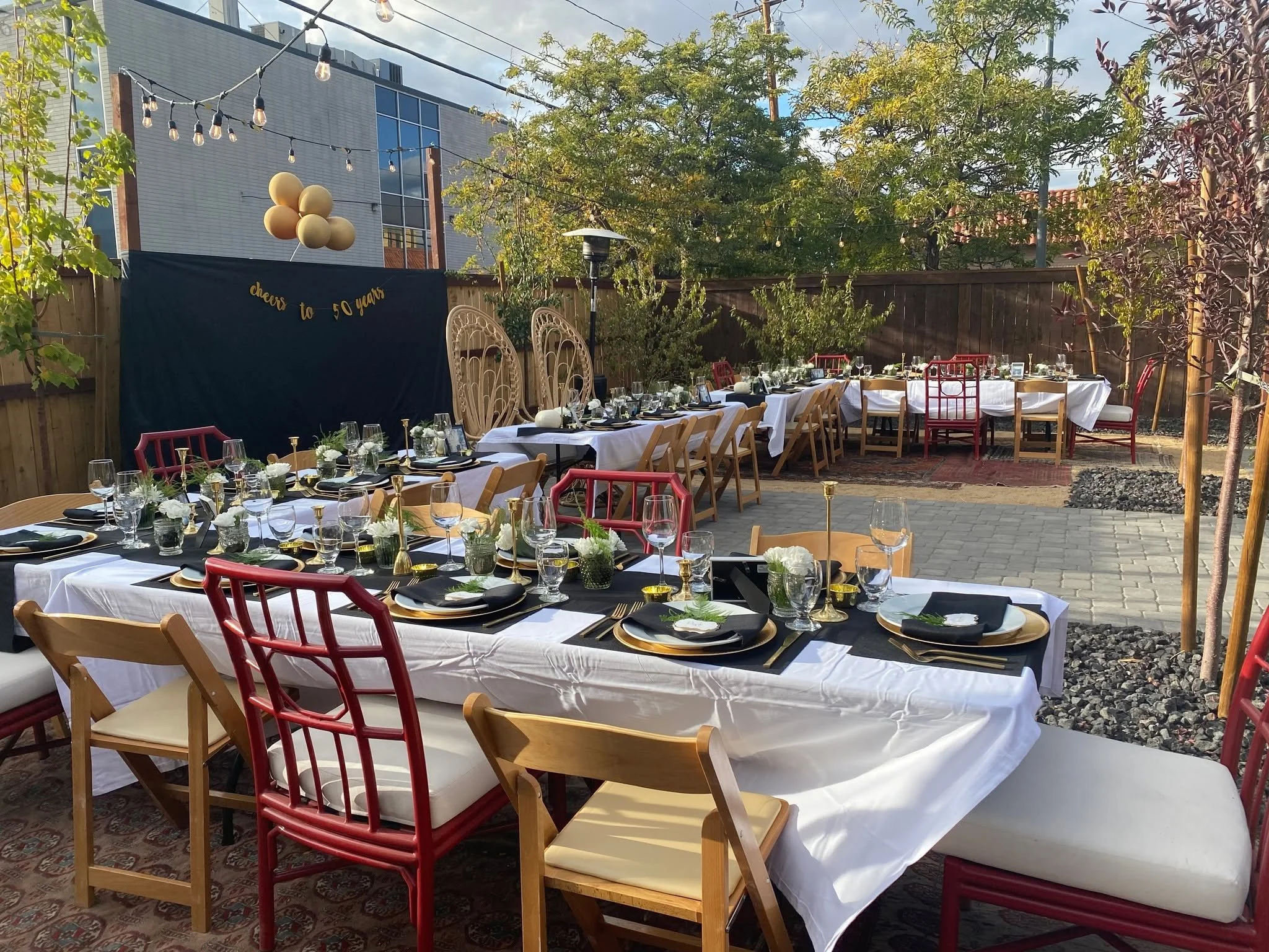 Outdoor celebration setup with long banquet tables covered in white tablecloths, decorated with floral centerpieces and glassware. Wooden and colorful chairs surround the tables. Decorative balloons, string lights, and a backdrop with a 
