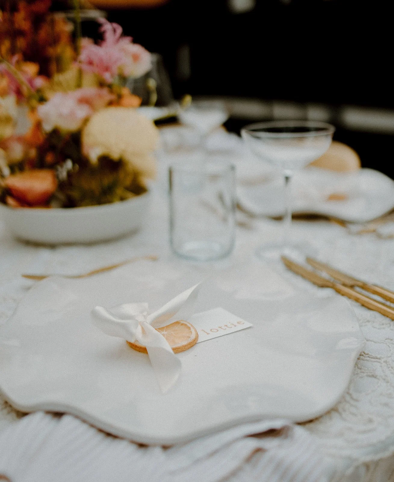 A table set for a celebration with a white tablecloth, a white cake with ribbon decoration, and a small tag with the word 'lotte'. There are empty glassware and floral arrangements in the background.