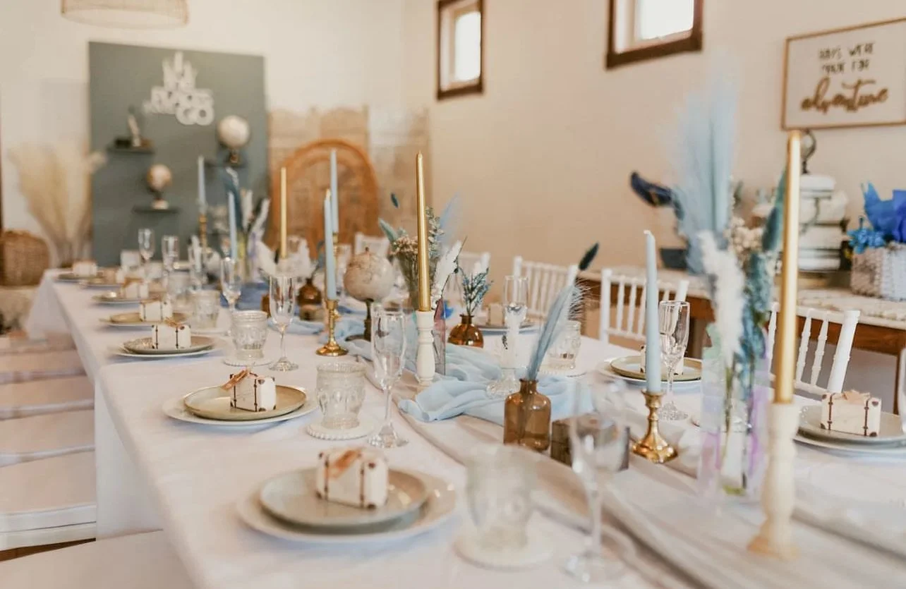 A decorated dining table with candles, flowers, and dinnerware in a well-lit room with windows and wall art.