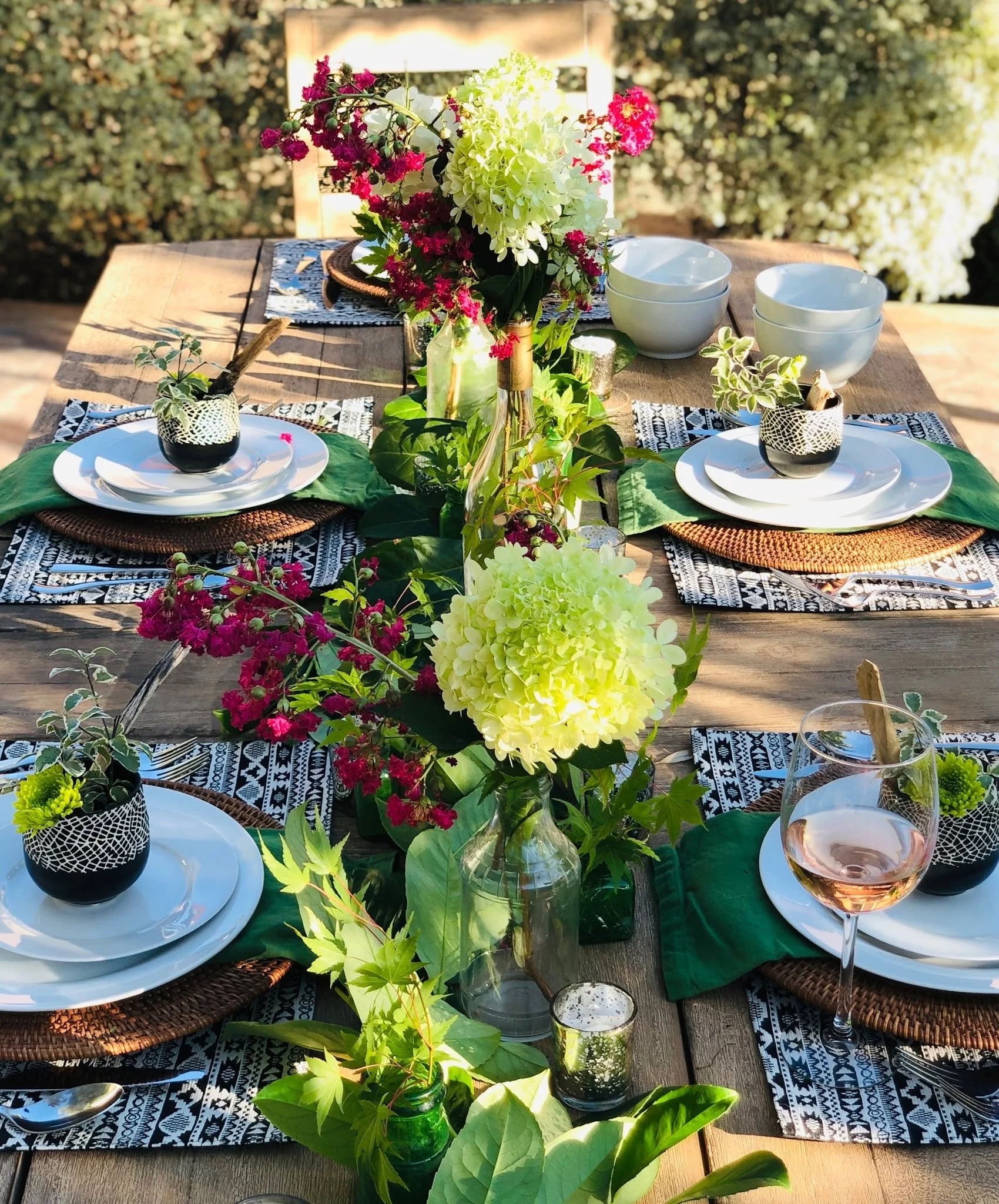 Table set for outdoor dining with white plates, black woven placemats, and decorated with lush green leaves, vibrant pink and white flowers in a glass vase, a glass of rosé wine, and small candles. The background features greenery and trees.