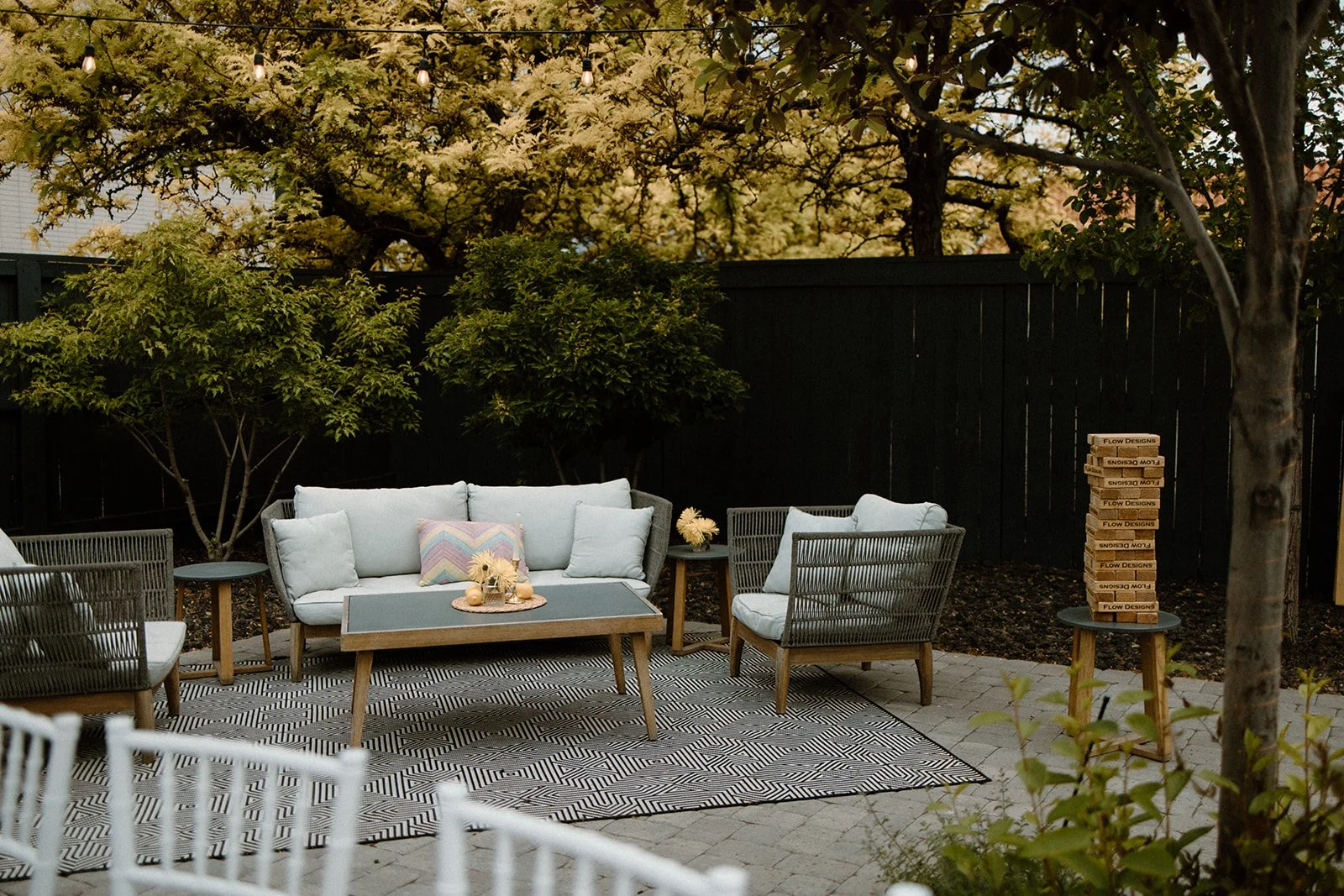 Outdoor patio with light-colored cushioned sofa and chairs, a rectangular coffee table with decorative items, two side tables with flower arrangements, a patterned outdoor rug, a black fence, green trees, and a stack of boxes on a small round table.